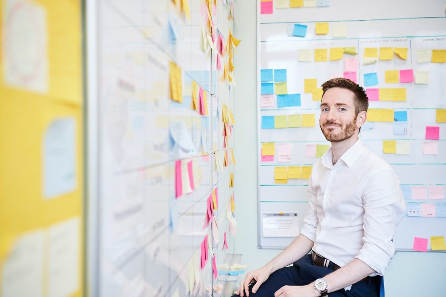 Man smiling to camera sitting in a room with whiteboards covering in sticky notes