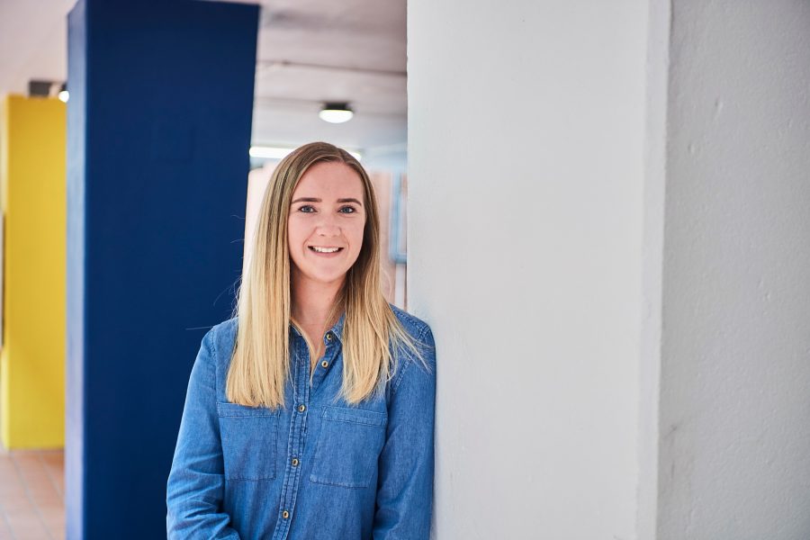 Young woman in denim shirt leaning against a wall in an office smiling to camera