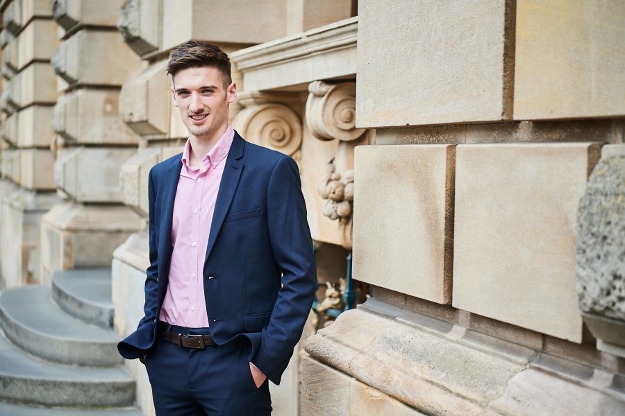 Young man looking into camera, smiling stood outside an architectural building