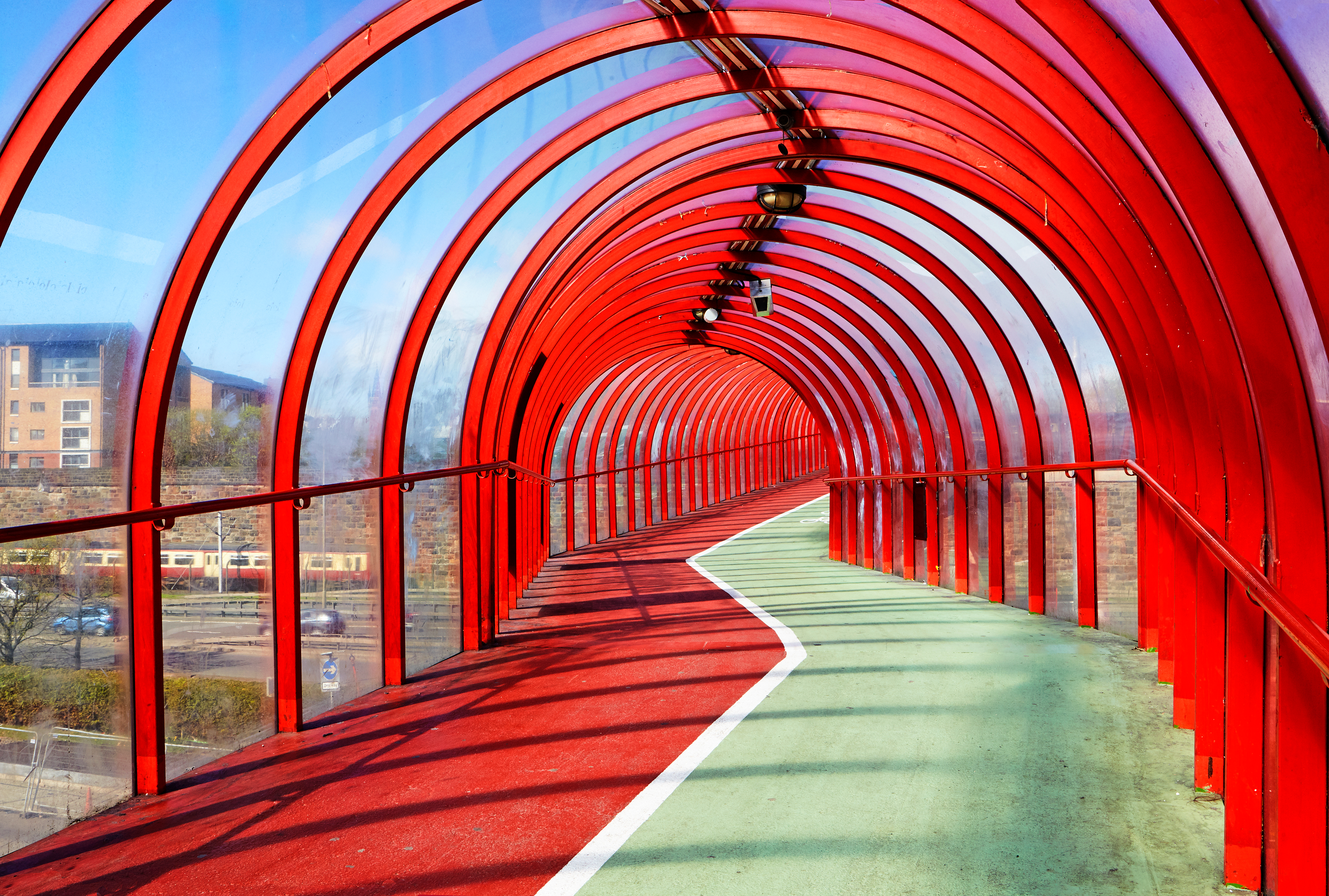 View from inside an enclosed tunnel consisting of red struts and clear panels that carries pedestrians and cyclists in Glasgow
