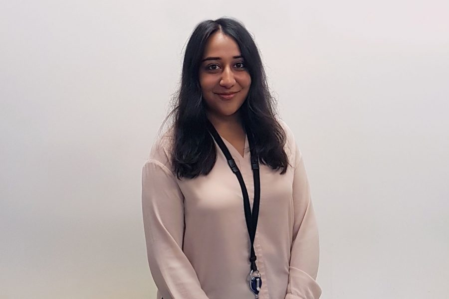 Woman with long dark hair and a work lanyard around her neck stood in front of a white wall, smiling at the camera